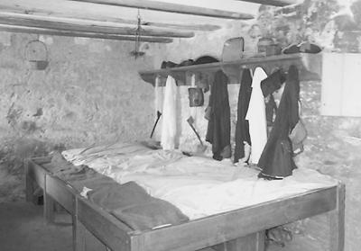 A black and white photograph of one of the interior rooms of Fort Matanzas in St. Augustine, Florida. The room is interpreted as soldier's quarters, with three wooden beds in the foreground lined up and touching each other. They are made up with white sheets and all have dark fabric at the head of the bed, maybe a burlap pillow. Above the foot of the bed is a wall shelf from which jackets are hung in a line parallel to the bed. On the shelf are various bags, shoes, and supplies.