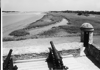 A black and white photograph of the Fort Matanzas gundeck overlooking the marshes of Fort Matanzas National Monument, which is controlled by the National Parks Service. Two cannons are on the gundeck in the foreground, pointing south out towards the waterway. To the right of the cannons, a sentry tower extends from the coquina wall of the gundeck. It is also made of coquina and is cylindrical in shape. 