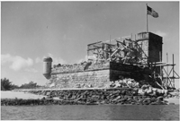 A black and white photograph of Fort Mantanzas National Monument in 1938. An American flag flies over the gun deck in the top right section of the image. Scaffolding covers two sides of the fort, the side facing the inlet and the second floor wall on the gun deck.