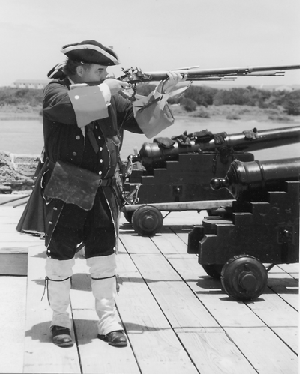 A black and white photograph of a historical reenactor portraying a Spanish soldier. He is a white man of strong build, with dark hair. He wears a tricorn hat, large jacket, pantaloons, and knee socks / canvas boot covers. He is pointing a period accurate musket to the right of the frame. In the background is the Matanzas gundeck with two cannons on it. The marsh and Rattlesnake Island are also visible.