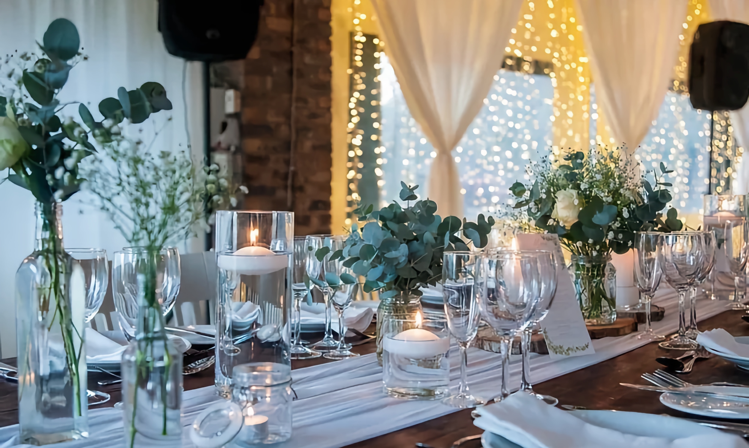 A long table set for an event, with crystal and glass, sparkle and greenery atop a dark wood table with white runners