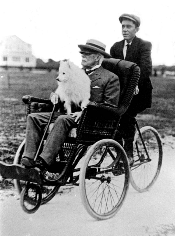 A black and white image of Henry Flagler on a foot-powered pedicab. He is sitting in a wicker chair with a set of wheels beneath him. A young man in a newsboy cap sits behind him, pedaling the device. Flagler is a distinguished old man with a suit, can, hat, and small white dog in his lap.