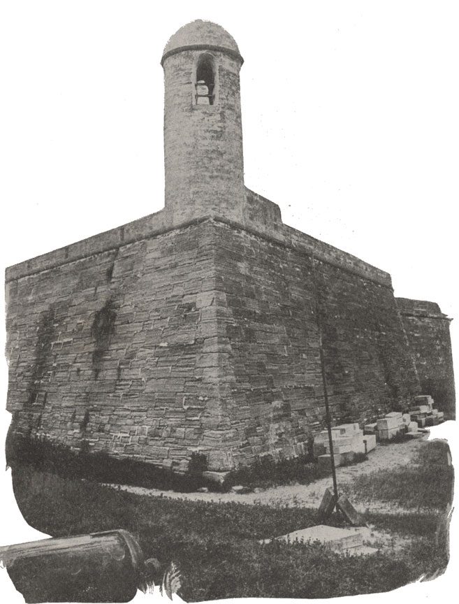 A black and white photograph of the north tower of the Castillo de San Marcos in St. Augustine, Florida. It is a coquina stone tower. There is a cannon on the ground in the foreground and the fort's 'moat' is filled with grass and damp mud.