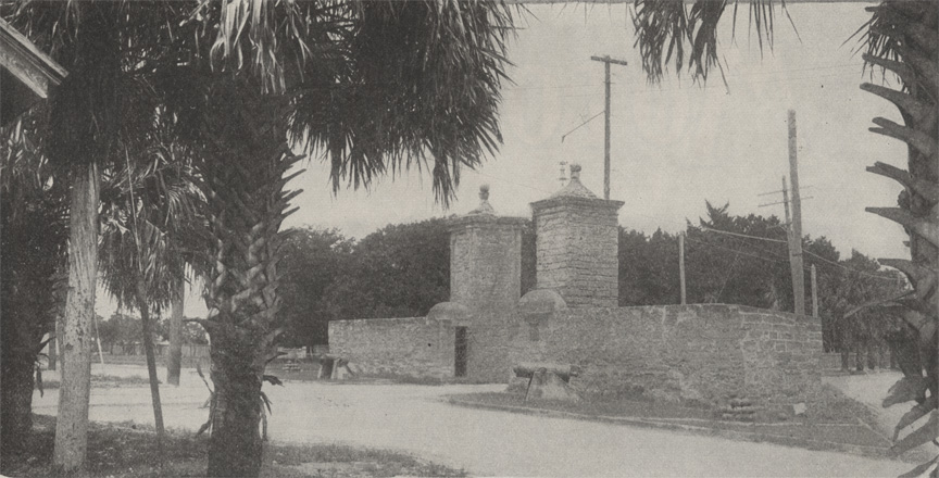 A black and white photograph of the St. Augustine City Gates from the north looking south. The gates are made of coquina stone. There are palm trees in the foreground.