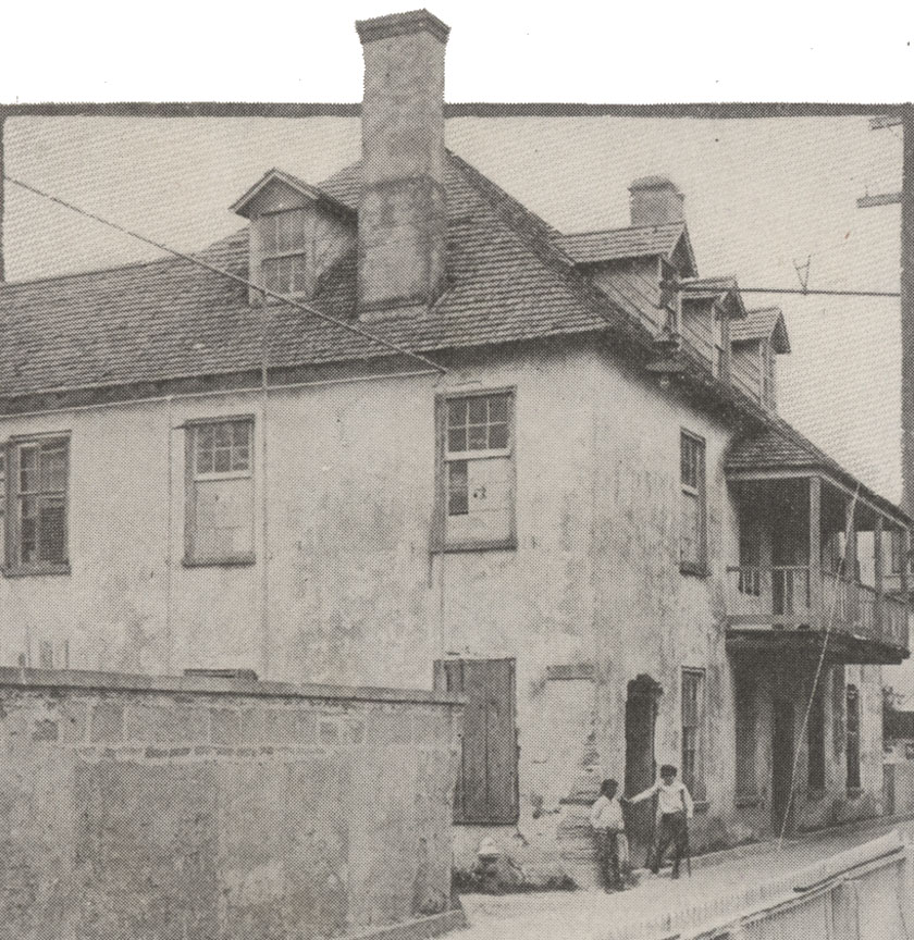 A black and white image of the Ximenez-Fatio House in St. Augustine, Florida, circa 1915. This image was taken on Aviles street, facing North. You can see the corner of the two-story coquina building. On the ground level, two boys lounge / pose for the camera. One has a bicycle between his legs.