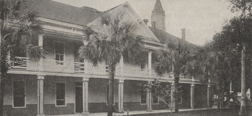 A black and white photograph of the Governor's House in St. Augustine, Florida. It is a three-story building with a wrap-around balcony on the second floor. The bell tower of the Cathedral can be seen in the background above the roof of the Governor's House, showing that the Cameraman was facing north.