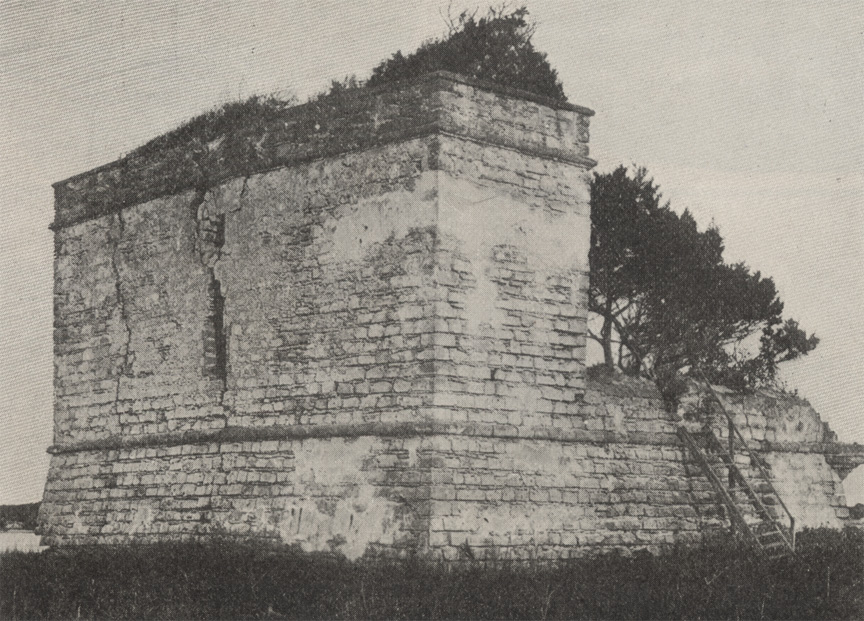 A black and white photograph of Fort Matanzas National Monument as it appeared circa 1915 -- its coquina stone structure is crumbling and its two gun decks are overgrown with marsh shrubbery. In St. Augustine, Florida.