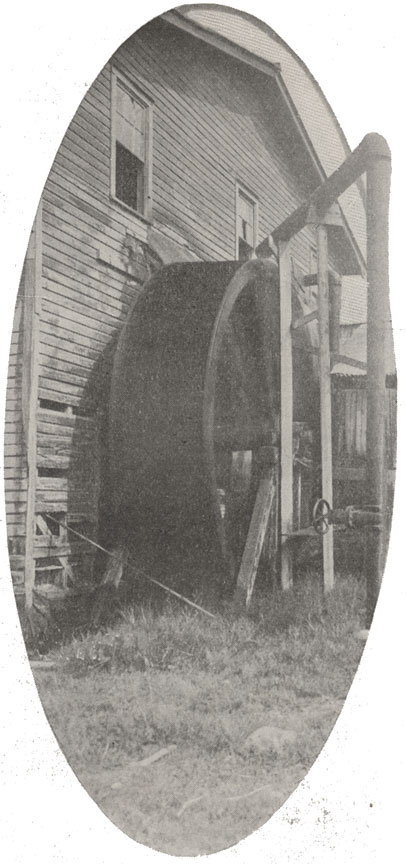 An ovular black and white photograph of a wooden two story house with a mill wheel on one wall.
