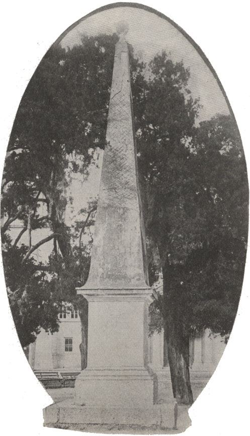 A black and white photograph of the constitutional monument in St. Augustine, Florida. It is a stone obelisk. There are old red cedar trees in the back ground. This image has an ovular frame.