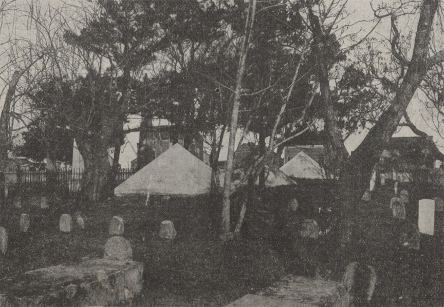 A black and white photograph of the National Cemetery in St. Augustine, Florida. Gravestones and trees can be seen in the foreground, with three large coquina pyramids in the background.