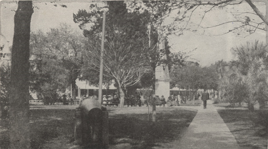 A black and white photograph of the Plaza de la Constitucion in St. Augustine, Florida, circa 1915. It shows people gathered on benches to sit and enjoy the day -- dozens in the background. A cannon sits in the foreground and you can also see the constitutional monument in the background.