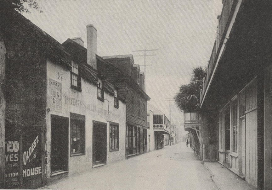 A black and white image of St. George Street in St. Augustine, Florida. Store fronts and museums like an empty sand street. Some houses have balconies. Som palm trees peek into the street from between buildings.