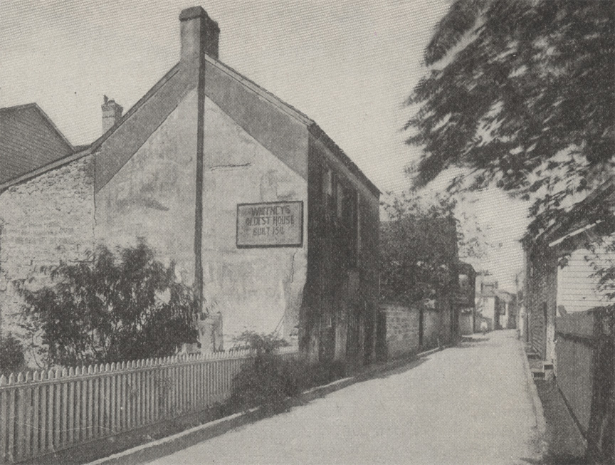 A black and white photograph of Whitney's Oldest House in St. Augustine, Florida. A dirt street with fences parallel to the street and historical houses lining both sides.