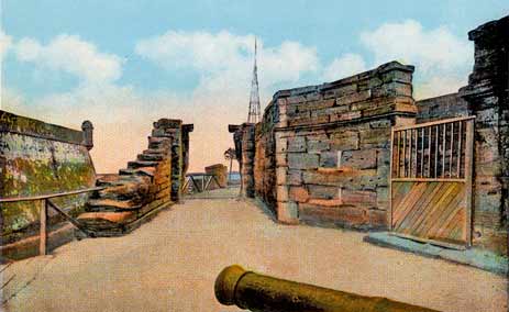 A color-tinted photograph of the First Approach to the Castillo de San Marcos, circa 1920s. The coquina walls of the lookout deck can be seen on the right, with a gate covering the stairs. A cannon's barrel can be seen peeking above the bottom edge of the photograph. The bastion of the fort can be seen in the background, in front of the Matanzas Bay. There is also some sort of triangular spire in the background.