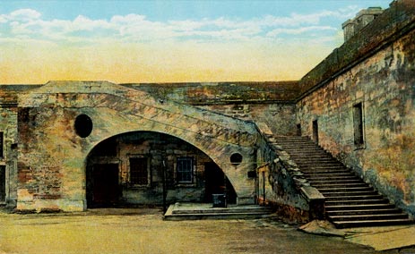 A color-tinted photograph of the inside of the Castillo de San Marcos (then Fort Marion), circa 1920s. It shows the coquina stairs to the gundeck on the right side of the image. To the left of the stairs is that peculiar arch, which leads to the public restrooms in present day. The floor of the fort is dirt and the sky is blue.