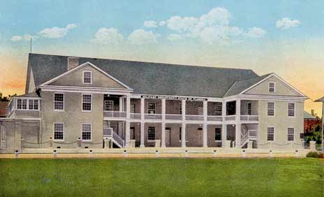 A color-tinted photograph (taken in the 1920s) of the St. Francis Barracks in St. Augustine, Florida. It is a building with three stories. The center section is recessed into a patio on the ground level and a white-railed balcony on the second level. Its walls are beige and its roof is grey.