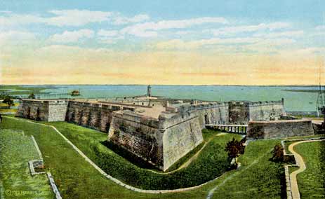 A color-tinted photograph of the Castillo de San Marcos (then Fort Marion) around the 1920s. It is an aerial view that shows the green grass in the multi-layered moat, the coquina walls of the fort, and the Matanzas Bay and Atlantic Ocean in the distance.