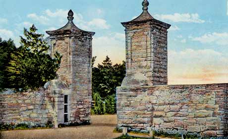 A color-tinted photograph of the St. Augustine City Gates in St. Augustine, Florida. They are two coquina stone pillars that extend to walls on either side, the walls being about half the height of the pillars. The pillars have triangular tops that have spheres on top of their points. The road that goes between them is dirt and the sky is tinted blue with a yellow glow on the horizon. Circa 1920s.