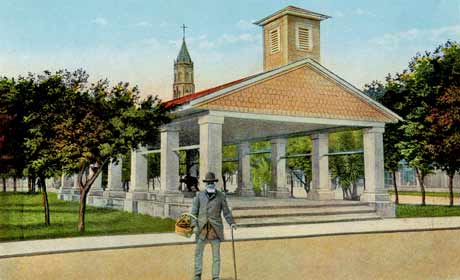 A color-tinted photograph of the Old Slave Market in St. Augustine, Florida. Also called the Public Market. It is a open-air pavillion with a triangular roof that has a structure to hold a bell that faces the Matanzas Bay. Behind the market, the plaza has green grass and rows of short trees. A man stands in front of the market, leaning on a cane and holding a basket in the other hand.