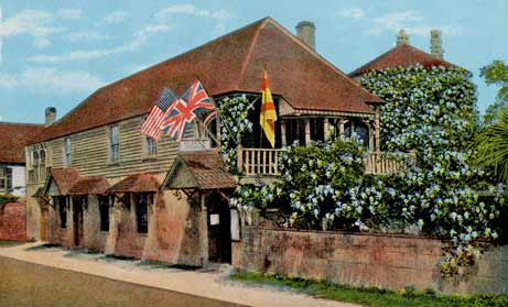 A color-tinted photograph of the Oldest House Museum in St. Augustine, Florida, circa 1920s. It is a long two-story house that runs parallel to St. Francis Street. Its first story is coquina stone and its second story is wood panel. Its roof is tall. The second story balcony flies three flags -- American, British, and Spanish. A large tree with blue flowers lines the wall to the right side of the image and climbs the building.