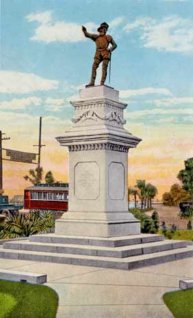 A color-tinted photograph (taken in the 1920s) of Anderson Circle in St. Augustine, Florida. In the center of the image, a statue of Juan Ponce de Leon pierces a slightly cloudy sky. Ponce is a bronze statue pointing to the left of the frame that stands on a white podium. There are three steps up to the podium from the ground. City buildings can be seen in the background.