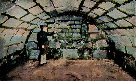 A color-tinted photograph of a room within the Castillo de San Marcos (then Fort Marion) circa 1920s. It is a room with an arched ceiling, made of bricks of coquina. A man stands in front of the back wall of this room, seemingly pointing to something on the wall. The dirt floor is tinted a pinkish brown. 
