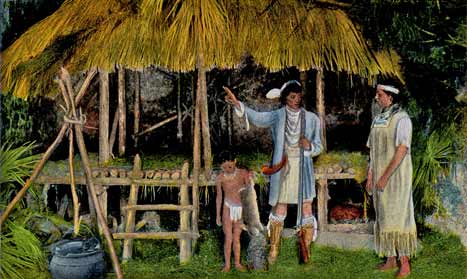 A color-tinted photograph of an interpretive display of Seminole life. Three figures stand on the rightside of the image in the foreground. The woman is the rightmost figure, with a man and a child to her left. The man points towards a thatched hut that sits almost like a porch, with a grass roof. On the left side of the image in the foreground, a pot sits beneath a teepee shaped fire spit.