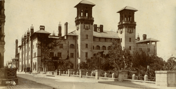 A sepia-toned image of the Alcazar Hotel in St. Augustine, Florida. It is a stone structure with two towers on its North-facing side and at least four floors. It spans about half of the block that it occupies. Circa 1888, its courtyard is full of tropical plants.