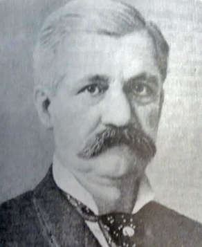 A black and white photograph portrait of Dan Harkness. He is a mustachioed older man with a somber gaze. He has dark gray hair that is very well groomed. He wears a tie with a pin.