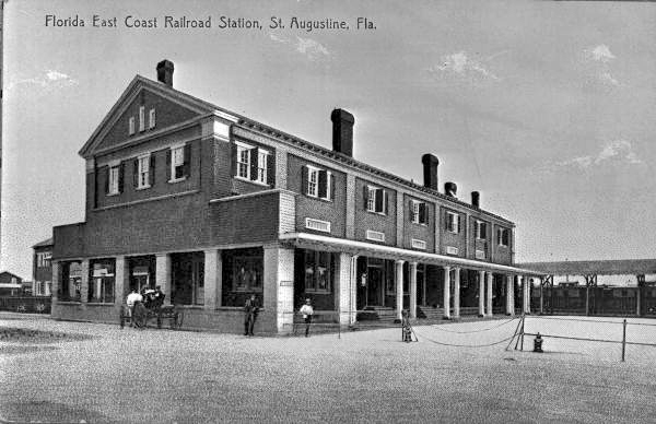 A black and white image of a three story building which was a train station in St. Augustine, Florida. It has five visible chimneys and the bottom level is a porch-type layout with recessed facilities under an awning. People linger outside the building on the ground level. The ground around the building is cleared out dirt.