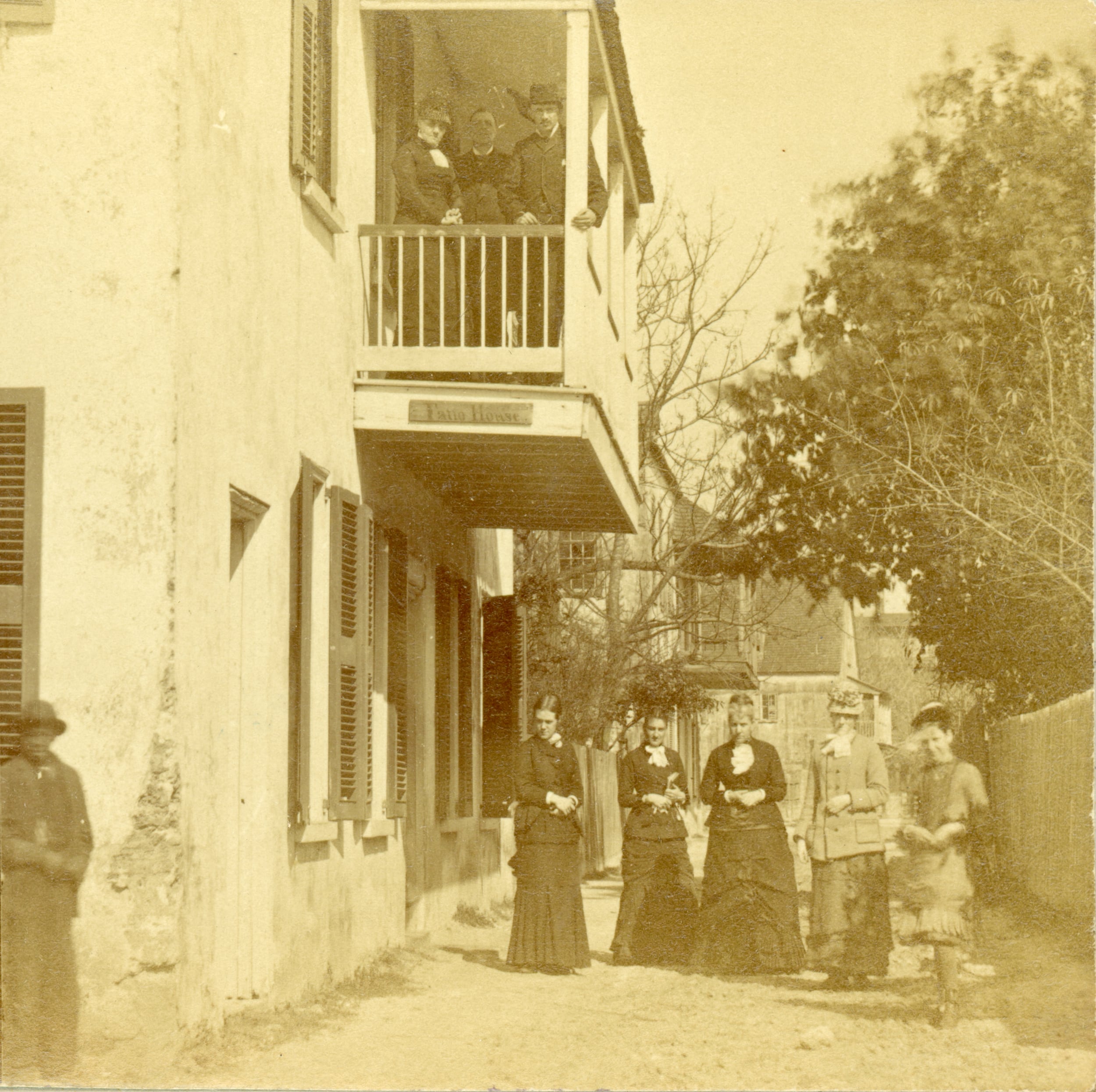 A historical image of the Ximenez-Fatio House in the 1880s, with white guests in the street and on the balcony, and a young black man at the corner of the house