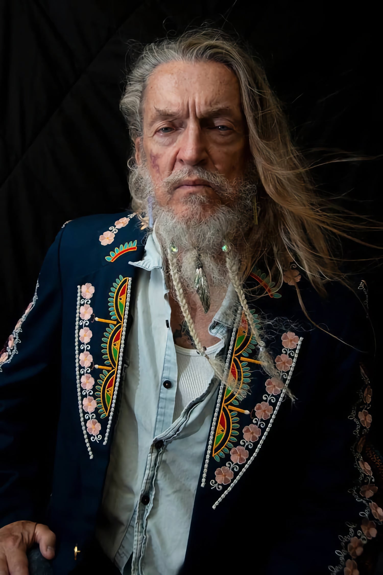 Ron Whitehead, an older man with long hair and a braided beard in front of a black background.