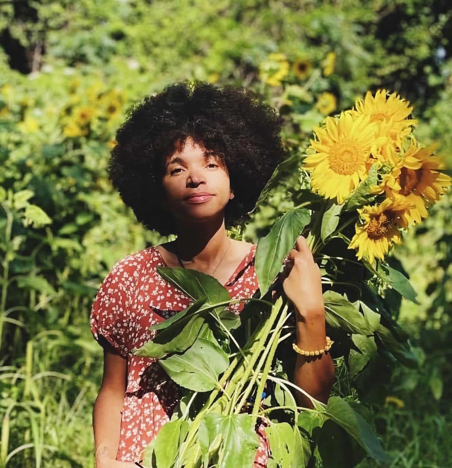Taylor Teachout, a young Black person, stands in front of green foliage holding a large sunflower.