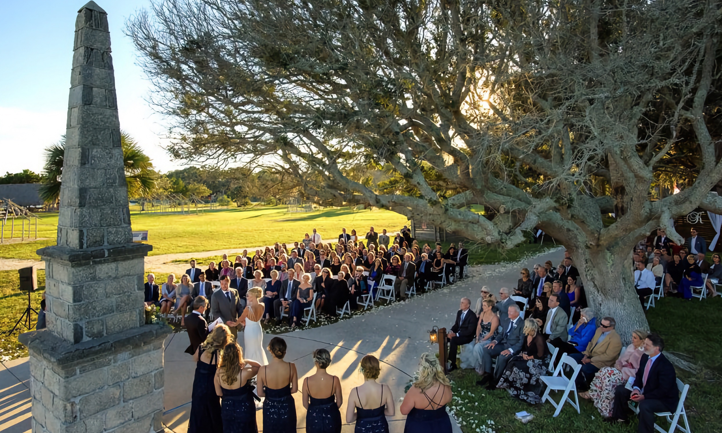 Beneath the branches of a live oak tree and a coquina obelisk, a couple ties the knot at the Fountain of Youth. The audience is seated in chairs on the grass around them.