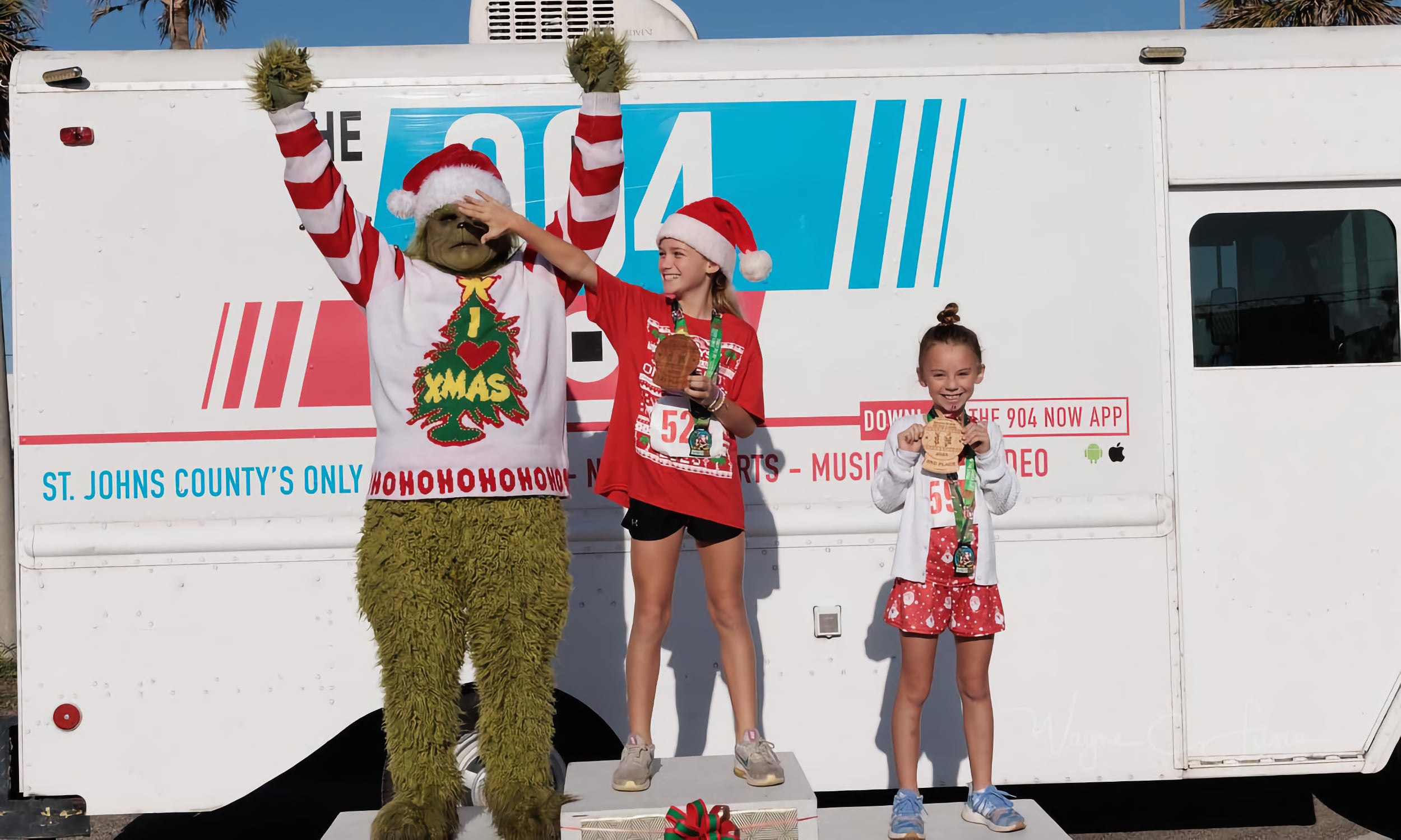 A person dressed as the Grinch celebrates with two girls showing their medals at the Santa Suits on the Loose 5K event in St. Augustine