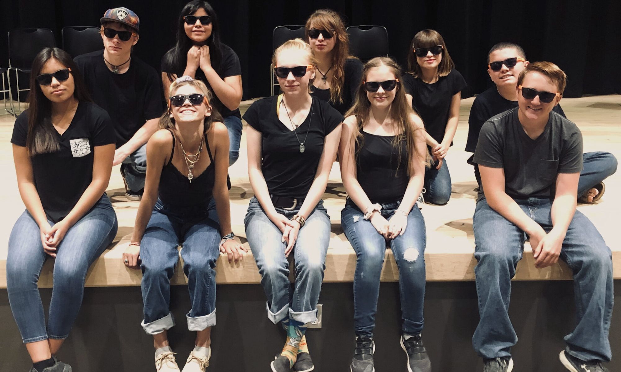 Teens in black t-shirts, jeans, and sunglasses sit on the stage.