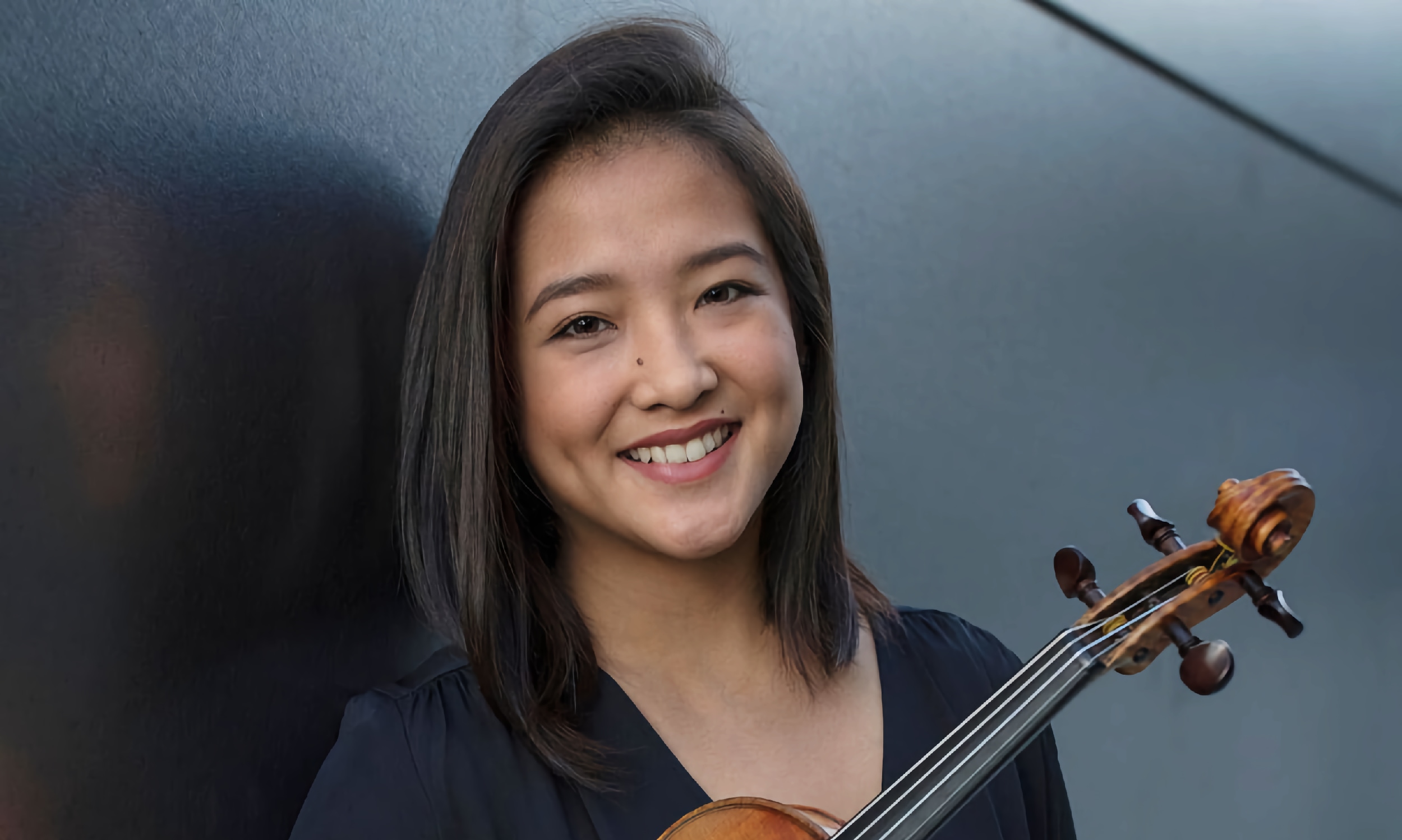 Gabriela Pena Kim, holding her violin and standing against a dark wall