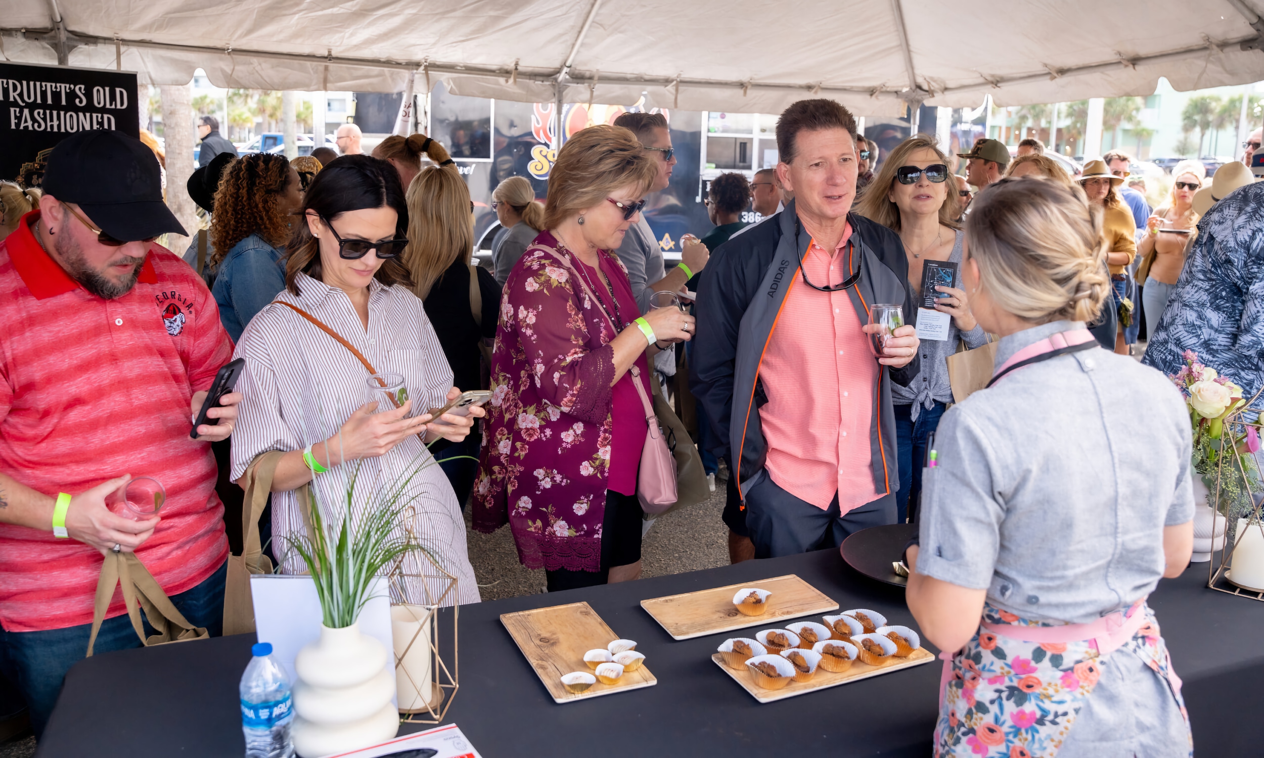 A woman stands at a table with sweet bites while people chat and taste the treats