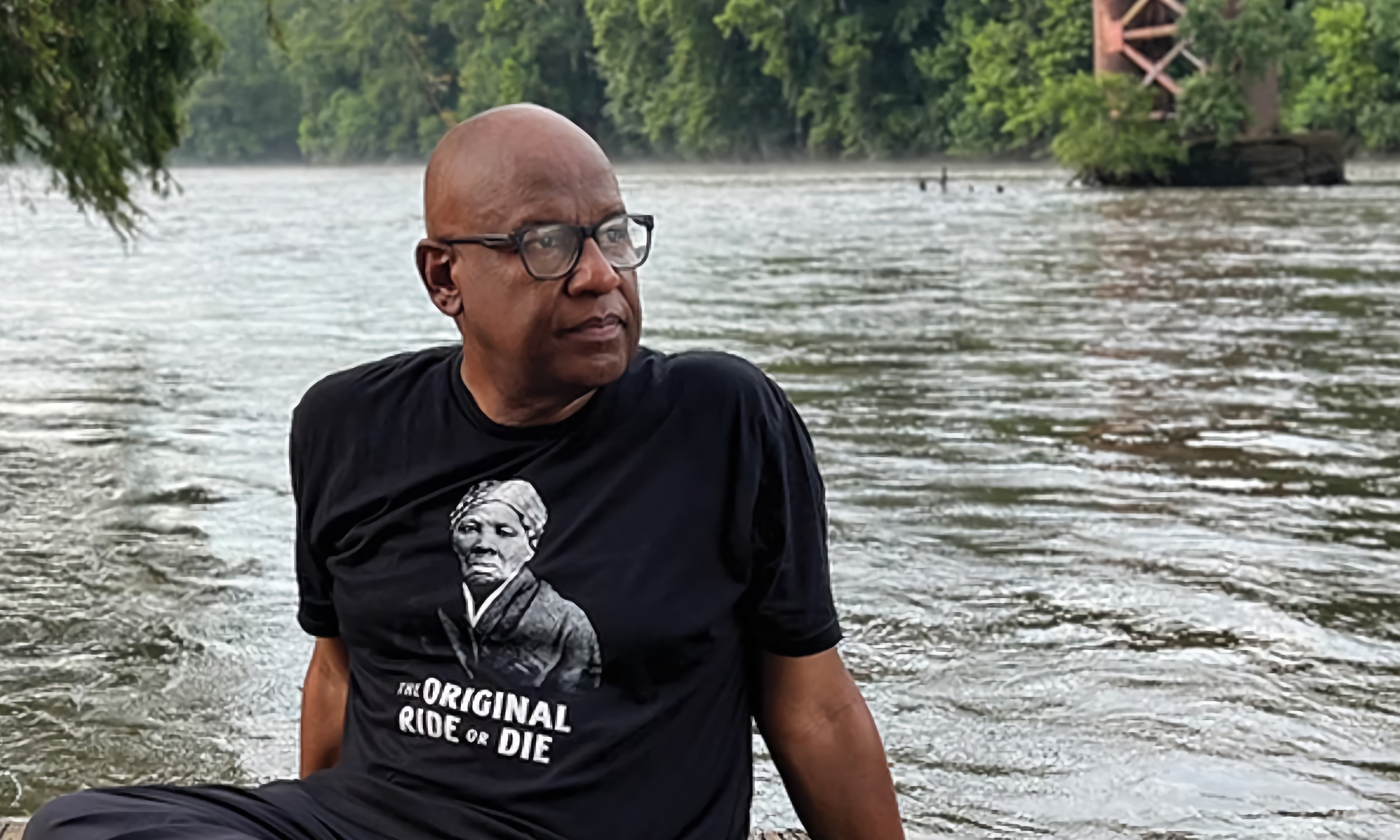Clennon L. King sitting next to a river, wearing a shirt with Harriet Tubman's image