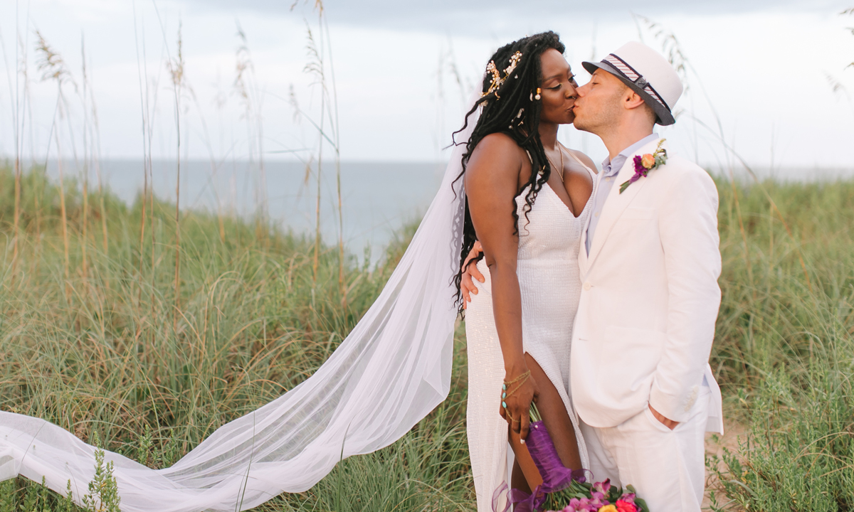 A bride and groom share a kiss in front of a dune on a beach in St. Johns County, with her veil draped across the grass