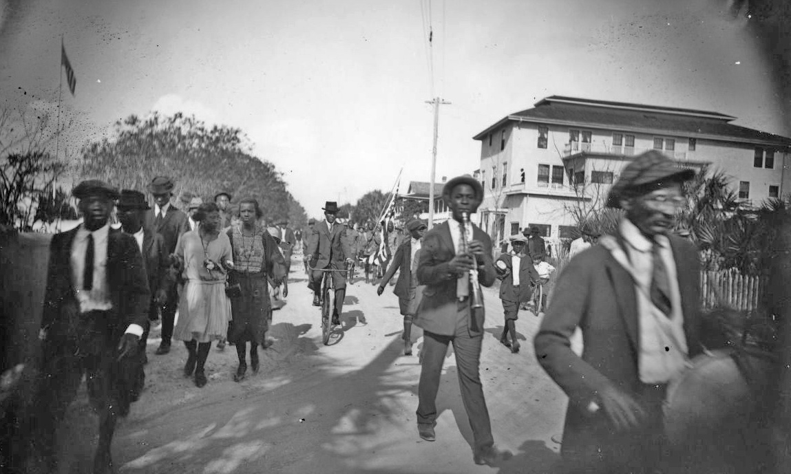 A historical image of a street in St. Augustine, Florida, Black residents of the city can be seen on parade, some playing instruments, some riding bikes. Image courtesy of Florida Memory.