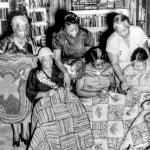 B&W a group of Black women display and discuss their quilts and crafts, which sit on their laps.