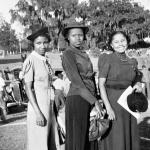 B&W, three young Black women pose in front of a line of cars.