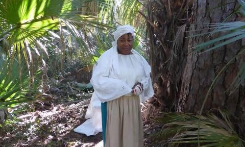 A black-American woman in the dress of the 1700s standing in a forest clearing