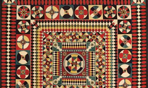 A detail view of an old quilt, predominately red, cream, and blue in color, and an intricate pattern that changes from the edges to the center