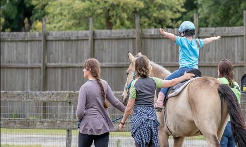 A child, atop a horse, arms akimbo, and flanked by three caregivers during equine therapy