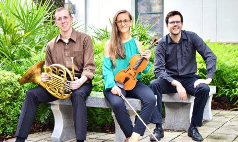 Three musicians, siting outside on a bench, two are holding instruments, a horn and a violin