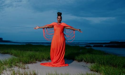  Mahoganee is a Gullahgeechee singer, songwriter, and poet. She is wearing a bright orange dress and spreads her arms out to embrace her fans. 