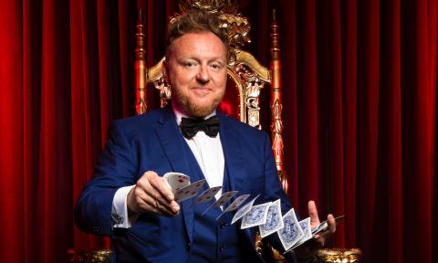 Magician Bill Abbot, in a blue tux, performs a card trick while sitting on a red and gold throne, in front of a red curtain