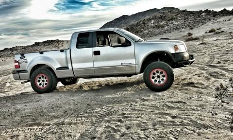 A silver Ford F-150 on a sandy desert hill. A cloudy sky and craggy mountains in the background